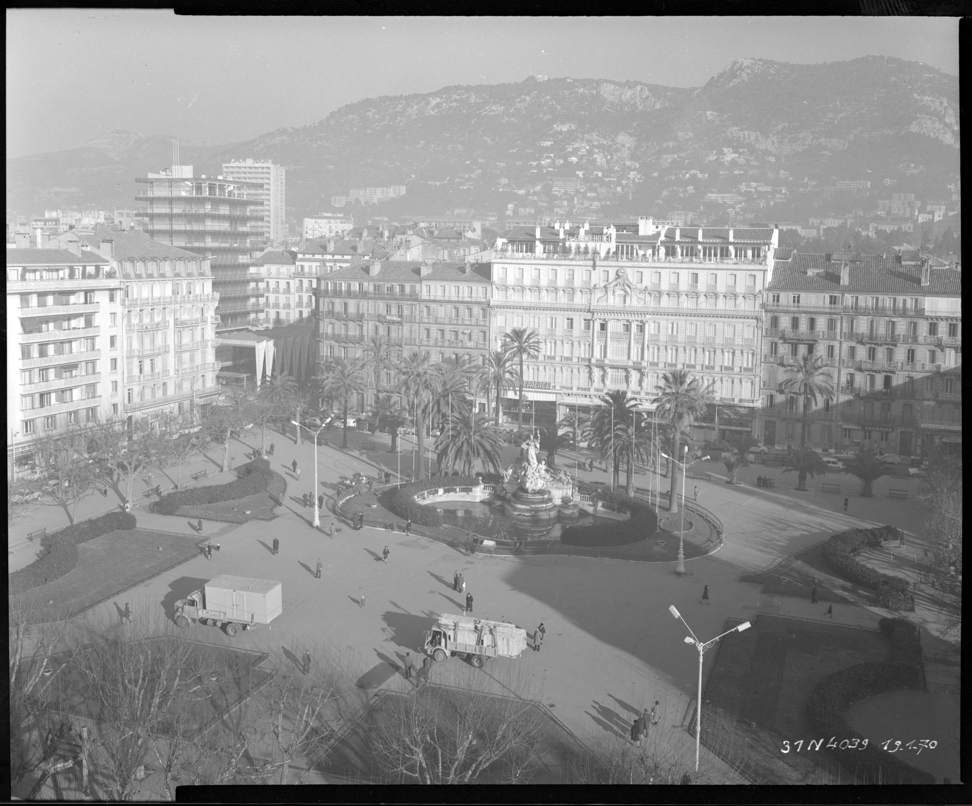 Vue générale en hauteur de côté, place, jardin, fontaine, immeubles, collines, deux camions sur la place, promeneurs, le grand hôtel